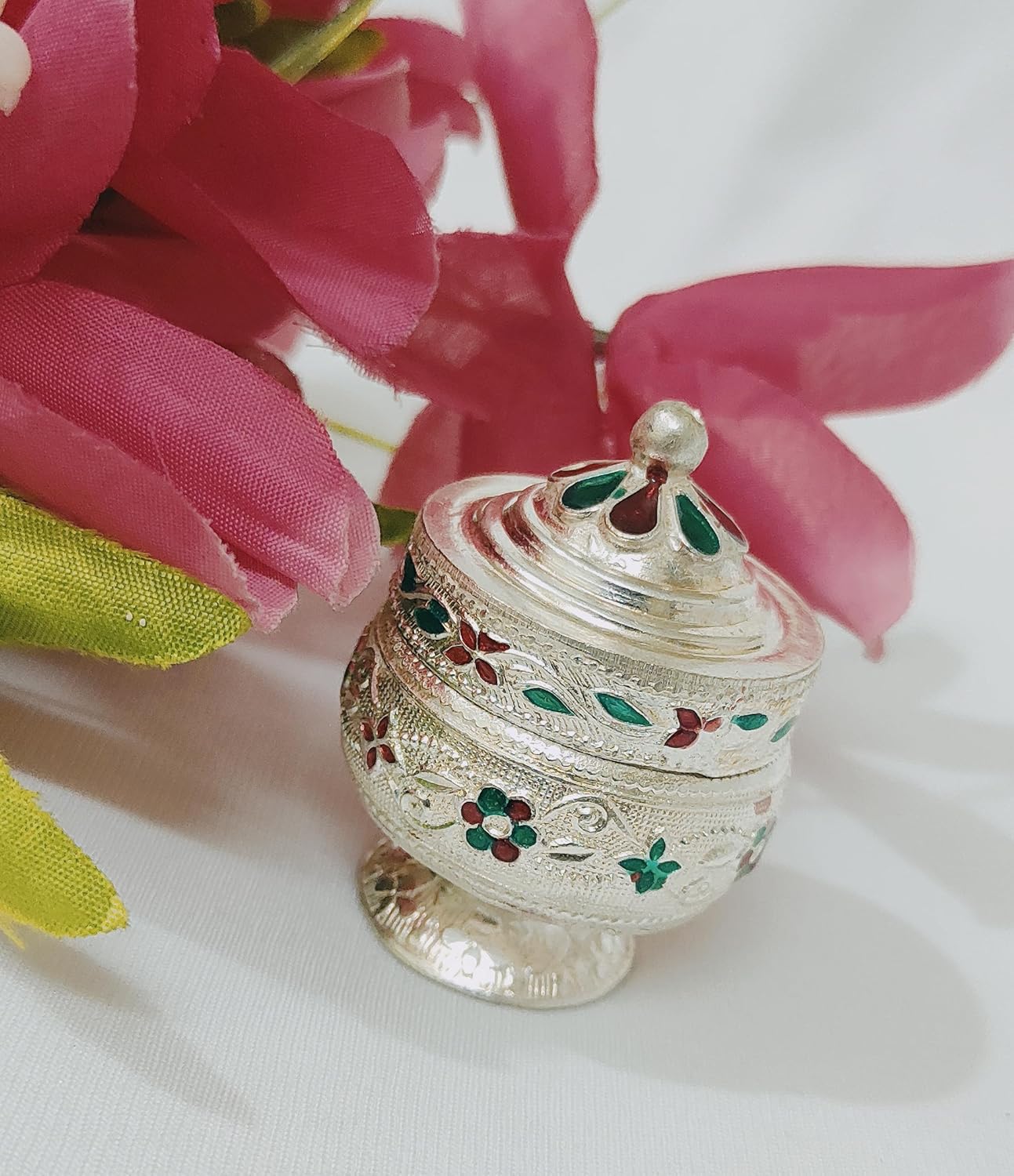 Decorative silver container with floral engravings on a white background with pink flowers.