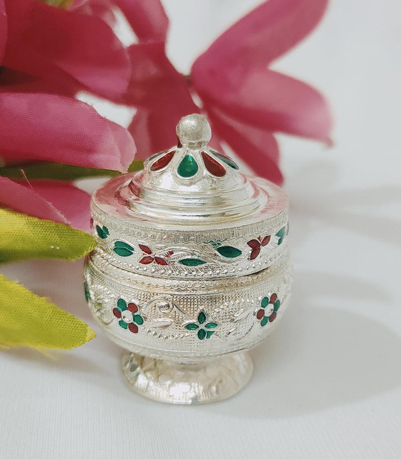 Decorative silver container with floral designs on a white surface with pink flowers in the background