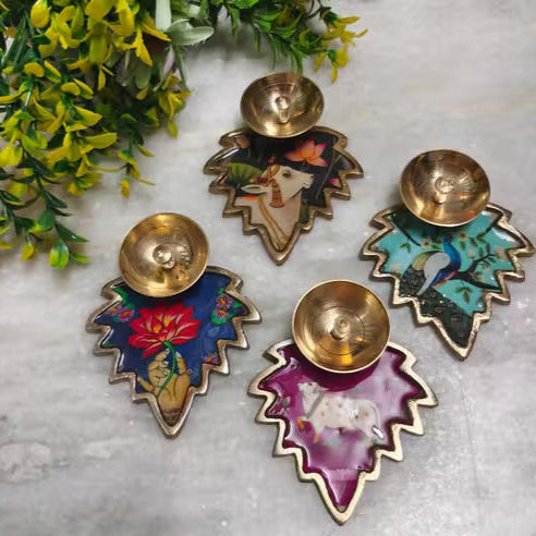 Decorative diya holders with colorful designs on a marble surface with greenery.